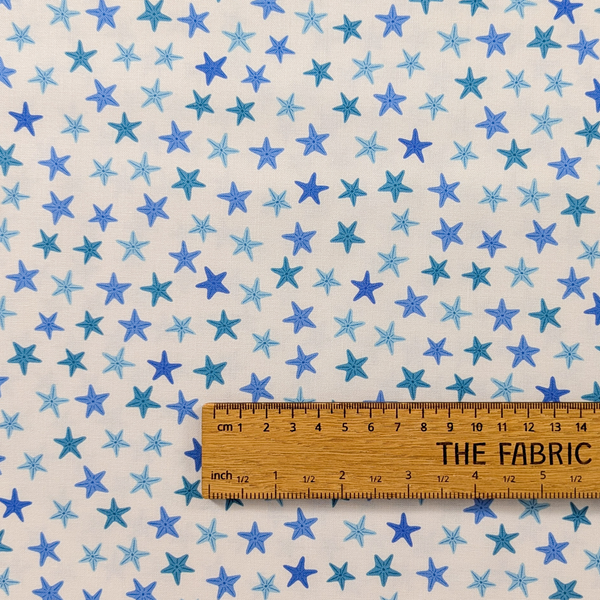 A repeating pattern of hand-drawn starfish in various shades of blue and teal on a warm off-white background. The ruler shows the scale of print.