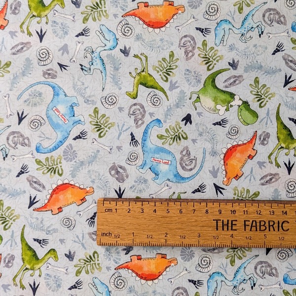 A repeating watercolor-style dinosaur pattern on a pale gray background, featuring blue, orange, green, and gray dinosaurs, bones, shells, and leafy motifs.  The ruler shows the scale of print.
