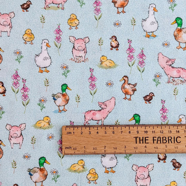A fabric showcasing cartoon-style pigs, ducks, and geese interspersed with delicate pink foxglove flowers and small white daisy-like flowers on a pale blue background with ruler for scale.