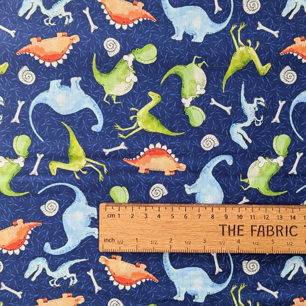 A repeating watercolor-style dinosaur pattern on a dark navy background, featuring green, light blue, and orange dinosaurs with white bones and spiral shell motifs.  The ruler shows the scale of print.