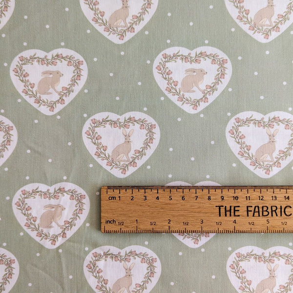 Repeating pattern of hearts with rabbits and floral wreaths on a sage green background with ruler for scale.