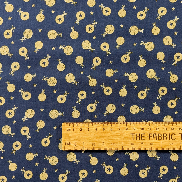 Navy blue fabric with a repeating pattern of small gold baubles with stars in the background.  A wooden ruler is laid across the bottom for scale.