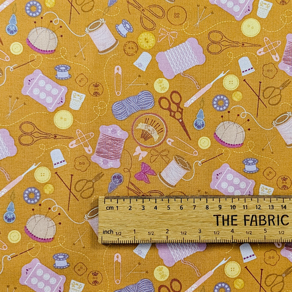 A repeating pattern of sewing and crafting tools on a warm tan background, including scissors, thread spools, pins, buttons, knitting needles, and yarn balls. The ruler shows the scale of print.