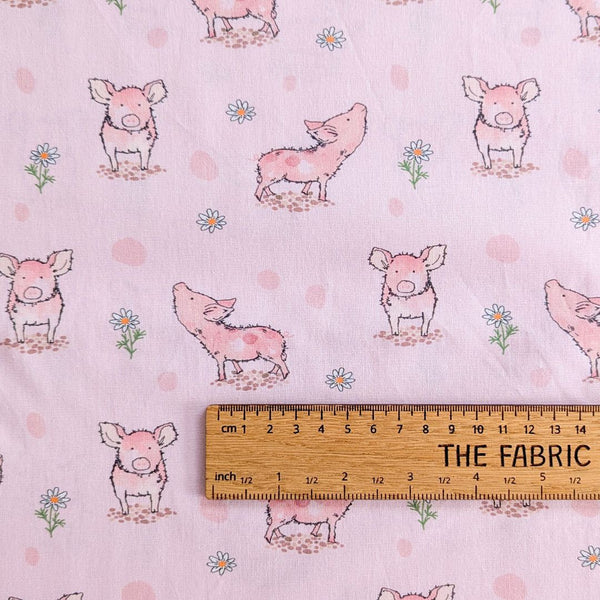 A fabric displaying adorable pink piglets in different poses, interspersed with small white daisy flowers and scattered with soft pink polka dots on a light pink background with ruler for scale.