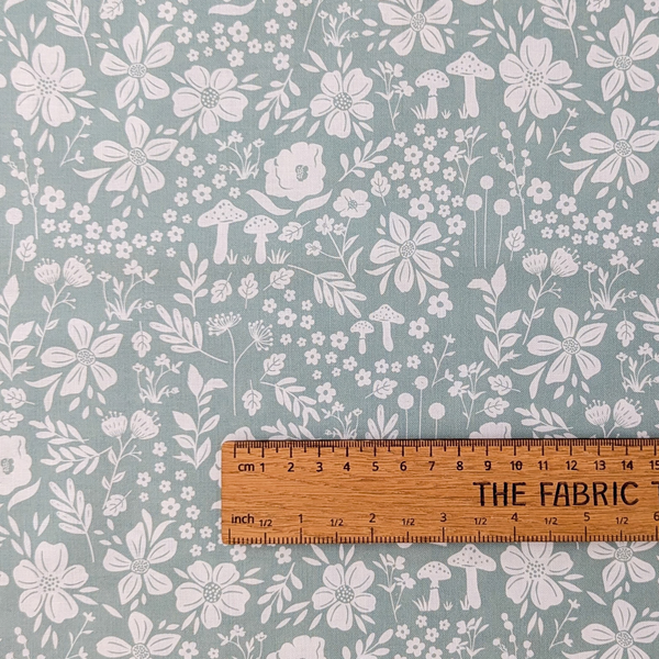 A repeating botanical pattern featuring white flowers, leaves, and small mushrooms on a muted sage green background.  The ruler shows the scale of print.