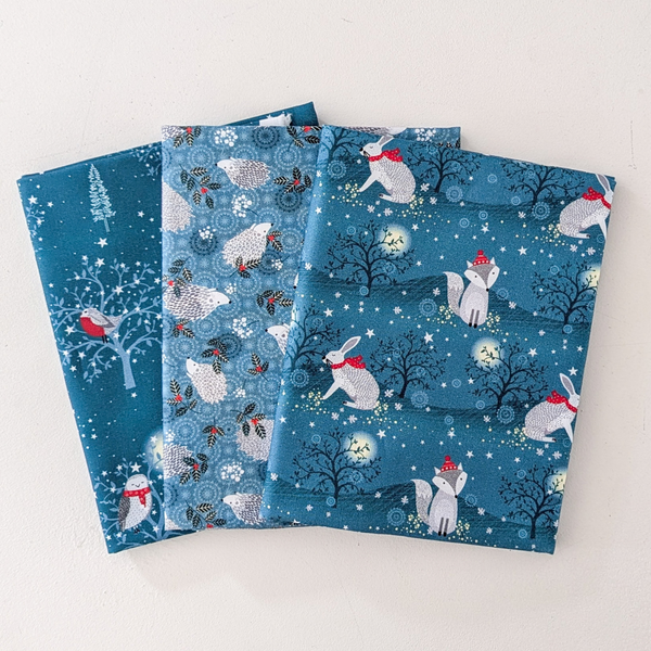 Three fabric swatches showing winter woodland patterns on a teal background: one with white rabbits and red scarves, one with white foxes and red hats, and one with white hedgehogs and red accents.