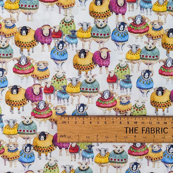 A repeating pattern of whimsical sheep wearing colorful patterned sweaters on a warm off-white background.  The ruler shows the scale of print.